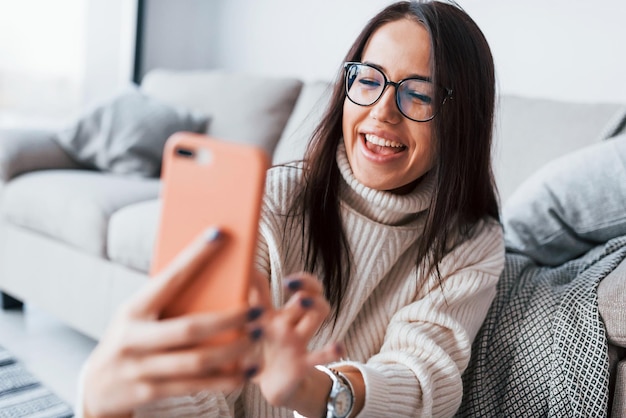 Faire des selfies. Jeune belle femme à lunettes assise seule à la maison avec un téléphone dans les mains.