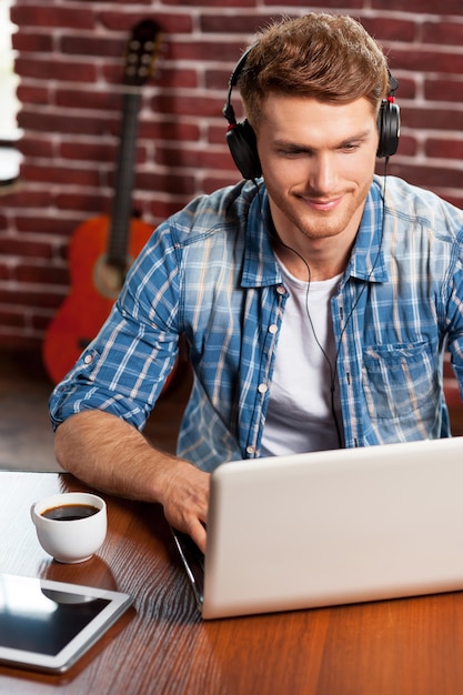 Faire un travail créatif. Vue de dessus du beau jeune homme travaillant sur ordinateur portable et souriant tandis que la guitare acoustique pose en arrière-plan