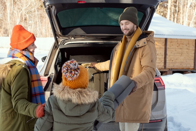 Famille active de trois personnes en vêtements d'hiver chauds mettant leur poussette dans le coffre en se tenant debout à l'arrière de la voiture, jeune homme prenant un tapis roulé