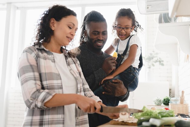 Une famille africaine heureuse et souriante de trois personnes, la mère cuisinant, faisant de la salade de légumes, tandis que le père et la fille l'aident à la cuisine.