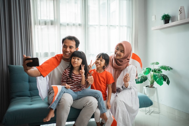 Une famille asiatique musulmane enthousiaste faisant un selfie et un appel vidéo en utilisant son téléphone à la maison tout en tenant le drapeau indonésien