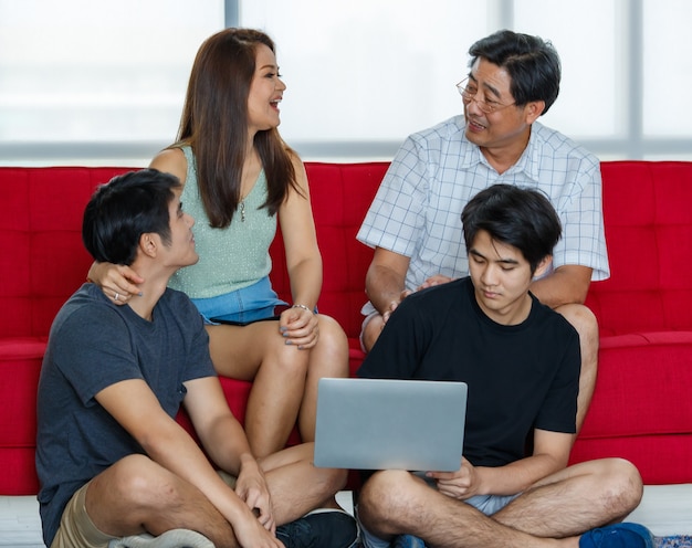 Famille asiatique de quatre membres composée de parents, de deux fils adultes passant joyeusement du temps ensemble pendant les vacances, utilisant un ordinateur portable dans un salon confortable à la maison. Présentation du concept de technologie moderne.