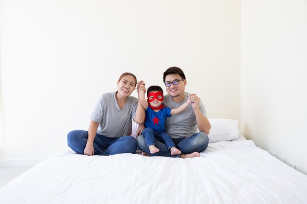 Famille asiatique et son fils portant un costume de super-héros la main et assis sur un lit blanc dans la chambre