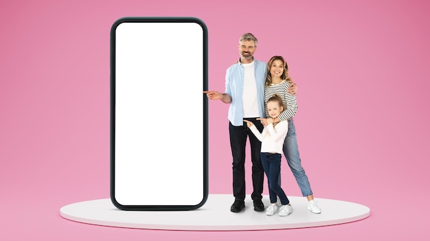 Une famille caucasienne adulte souriante montre des doigts sur un énorme smartphone avec un écran vierge approuvant l'annonce et