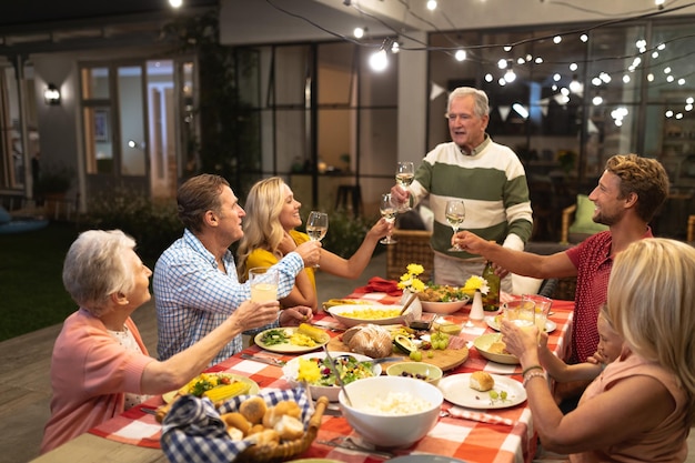 Famille caucasienne multigénérationnelle profitant de leur temps à la maison ensemble, assis près d'une table, parlant, se regardant et souriant, portant un toast