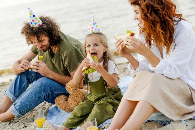 Famille avec des chapeaux de fête mangeant des sandwichs sur la plage Famille appréciant l'anniversaire de sa fille