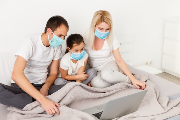 Famille dans des masques médicaux allongés sur un lit avec un ordinateur portable. Concept de regarder une vidéo, de travailler ensemble et de se mettre en quarantaine pendant une épidémie de coronavirus COVID-19