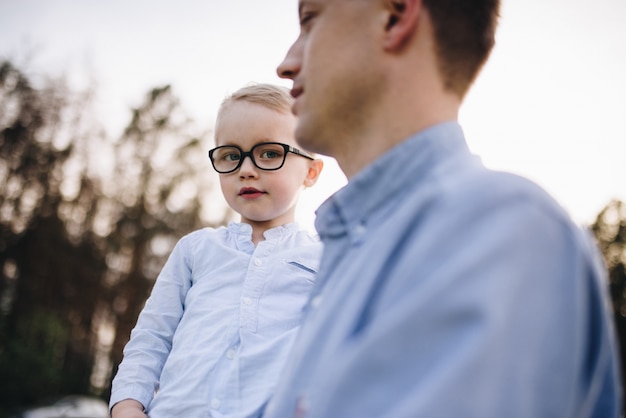 Famille Dans La Nature. Pique-nique En Forêt, Dans La Prairie. L'herbe Verte. Vêtements Bleus. Papa, Fils à Lunettes. Garçon Aux Cheveux Blonds. Joie. Les Parents Jouent Avec L'enfant. Ensemble. Panier Pique-nique. Nourriture, Couverture