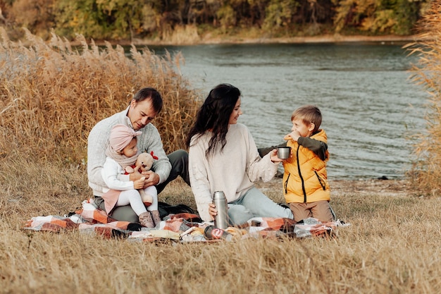 Famille dans le parc en automne. Femme dans un pull blanc. Enfants mignons avec des parents assis sur une couverture au bord du lac