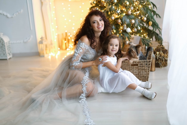 Une famille de deux personnes, maman et fille en robe blanche, est assise par terre sous l'arbre du Nouvel An.