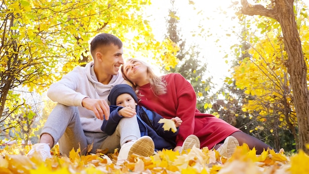 La famille du père, de la mère et du fils s'assoit sur des feuilles tombées sur fond d'arbres jaunes.