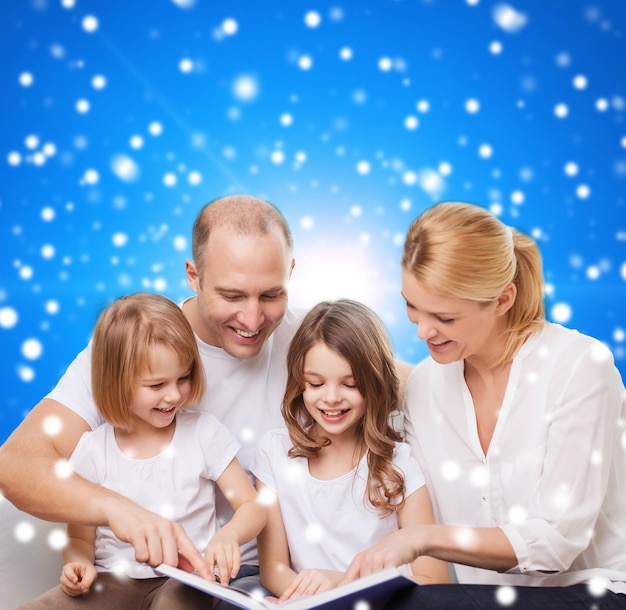 famille, enfance, vacances de noël et personnes - mère souriante, père et petites filles lisant un livre sur fond bleu enneigé