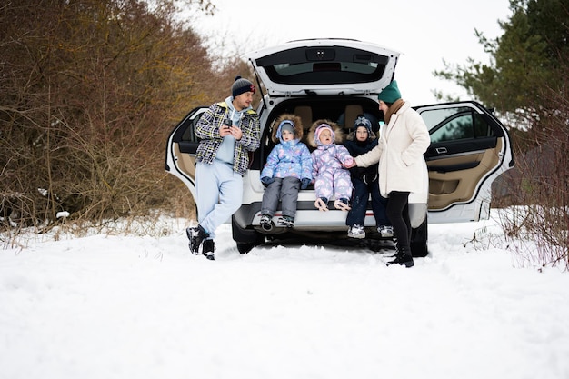 Une famille avec des enfants s'assoit sur une voiture suv avec un coffre ouvert dans la forêt d'hiver