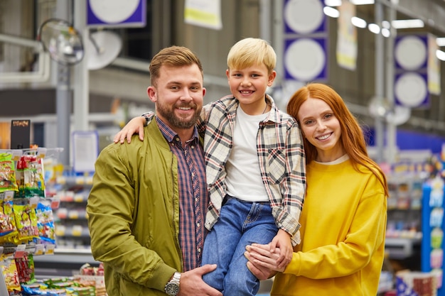 Famille avec fils au supermarché, les jeunes parents tiennent mignon enfant garçon dans les mains et sourire, étagères avec des produits