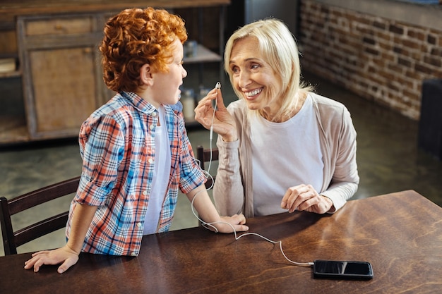 Famille harmonieuse. Mise au point sélective sur une femme âgée joyeuse qui sourit largement tout en mettant des écouteurs et en écoutant son petit-enfant aux cheveux bouclés lui parler.