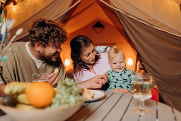 Une famille heureuse avec un adorable bébé dîne et passe du temps ensemble dans le glamping le soir d'été près d'un feu de joie confortable Tente de camping de luxe pour les loisirs de plein air et les loisirs Concept de style de vie