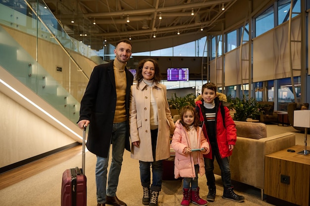 Famille heureuse appréciant le voyage ensemble, se tient avec des bagages dans le hall du terminal de départ de l'aéroport, attendant l'embarquement, sur le fond du panneau d'information avec l'horaire des vols