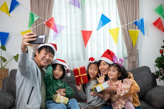 Famille heureuse asiatique avec fille en chapeaux de Père Noël réunis et appel vidéo à l'heure de Noël et profitez d'une boîte de cadeaux