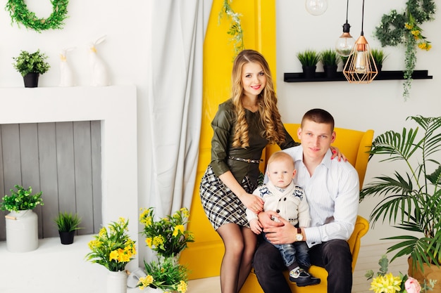 Famille heureuse, assis sur un fauteuil jaune chic, autour d'eux une cheminée, des pots de fleurs, des portes jaunes avec un rideau