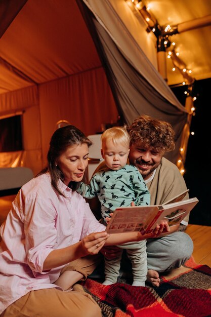 Famille heureuse avec un beau bébé jouant et lisant un livre ensemble dans le glamping le soir d'été près d'un feu de joie confortable Tente de camping de luxe pour les loisirs de plein air et les loisirs Concept de style de vie