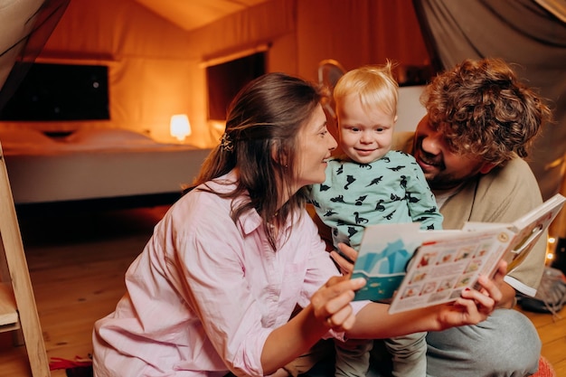 Famille heureuse avec beau bébé jouant et lisant un livre ensemble dans le glamping le soir d'été Tente de camping de luxe pour les loisirs de plein air et les loisirs Concept de style de vie