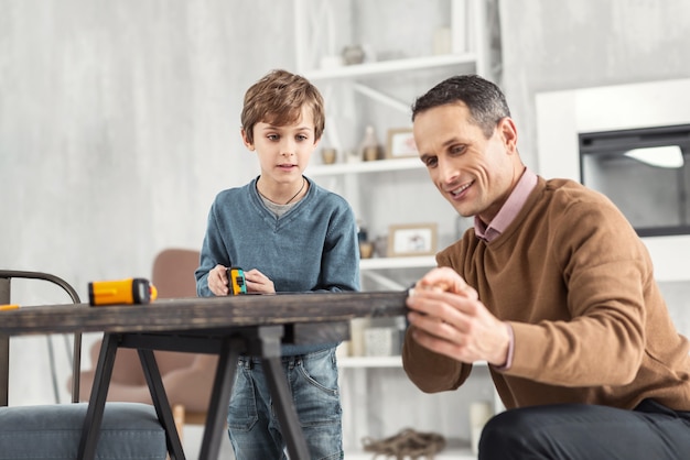 Famille heureuse. Beau contenu petit garçon blond tenant un ruban à mesurer et mesurant la table et son papa aidant à aider