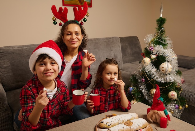 Famille heureuse, belle maman avec son fils et sa fille tenant de la guimauve et regardant la caméra assis à table avec du pain Stollen allemand fraîchement cuit, célébrant Joyeux Noël en cercle familial