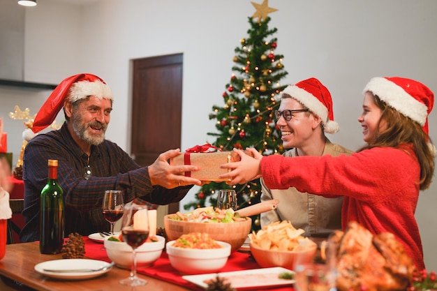 Famille heureuse célébrant le dîner de Noël à la maison, petite-fille avec boîte-cadeau donnée au grand-père.