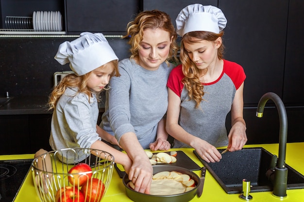 Famille heureuse dans la cuisine. Mère et deux enfants préparent la pâte, font cuire la tarte aux pommes. Maman et ses filles cuisinent des aliments sains à la maison et s'amusent. Ménage, aide au travail d'équipe, concept de maternité