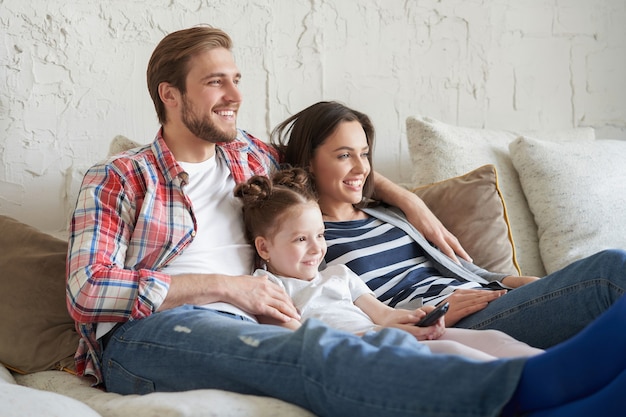 Famille heureuse avec un enfant assis sur un canapé en train de regarder la télévision, de jeunes parents embrassant leur fille se relaxant sur un canapé ensemble.