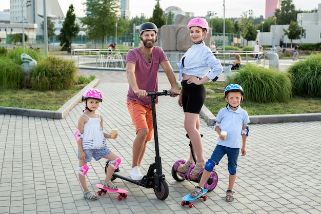 Famille heureuse avec enfants à cheval sur Segway, scooter électrique et planches à roulettes dans le parc en été, enfants mangeant de la crème glacée.