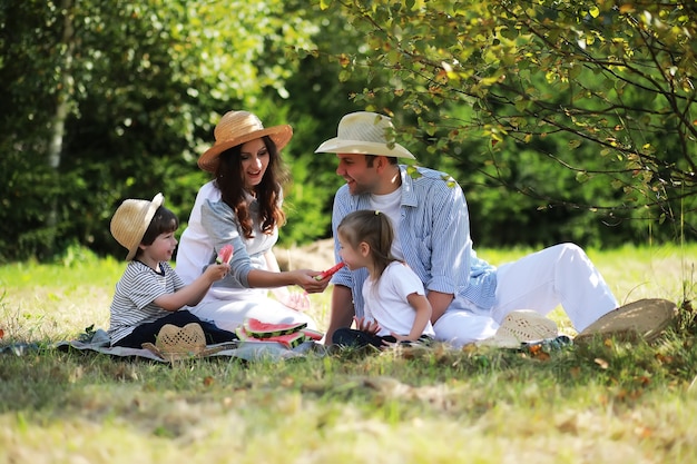 Famille heureuse avec enfants pique-niquant dans le parc, parents avec enfants assis sur l'herbe du jardin et mangeant de la pastèque à l'extérieur