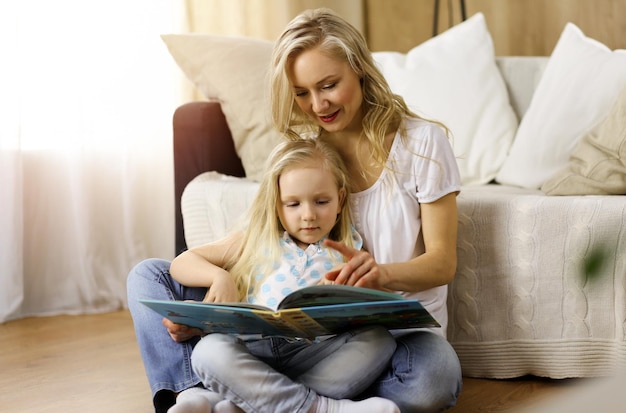 Famille heureuse. Jeune mère blonde lisant un livre à sa jolie fille assise sur un plancher en bois dans une pièce ensoleillée. Notion de maternité.