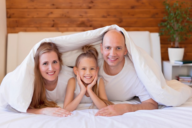 Famille heureuse, maman, papa et fille au lit à la maison dans la chambre sous les couvertures