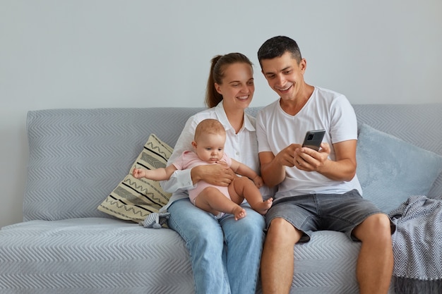 Une famille heureuse posant dans le salon sur un canapé, une mère tenant une petite fille, un père montrant un contenu intéressant via un téléphone portable, des gens souriants naviguant ensemble sur Internet.