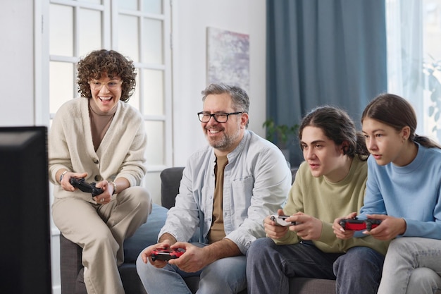 Une famille heureuse de quatre personnes jouant ensemble à une console de jeu tout en étant assise sur le canapé dans la chambre à la maison