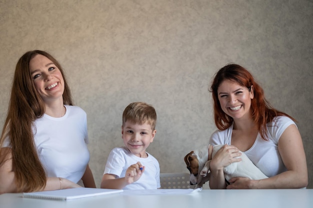 Une famille heureuse reste à la maison Deux femmes aident le garçon à faire ses devoirs scolaires Couple de lesbiennes assis à la table avec leur fils et un chiot joyeux Mariage homosexuel avec un enfant Apprentissage à distance