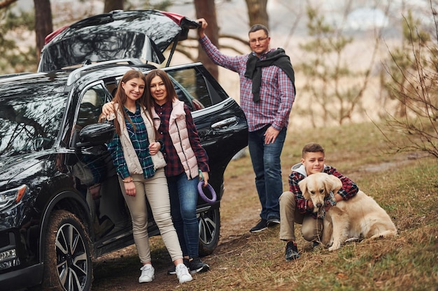 Une famille heureuse s'amuse avec son chien près d'une voiture moderne à l'extérieur dans la forêt