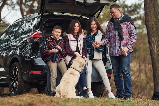 Une famille heureuse s'amuse avec son chien près d'une voiture moderne à l'extérieur dans la forêt