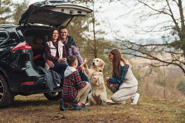 Une famille heureuse s'amuse avec son chien près d'une voiture moderne à l'extérieur dans la forêt