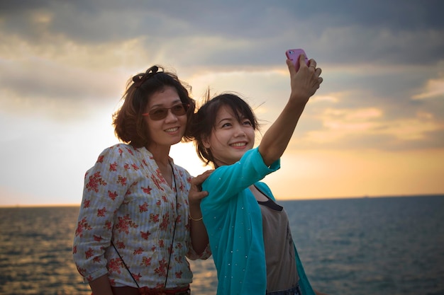 Une famille heureuse se fait un selfie avec son smartphone alors qu'elle se tient sur la plage contre un ciel nuageux au coucher du soleil.