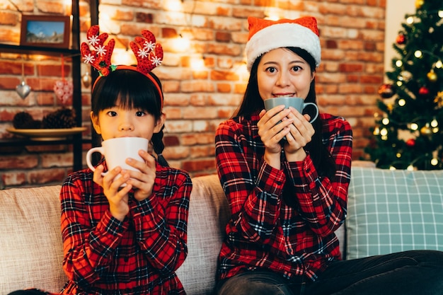 famille heureuse tenant une tasse à boire sur un canapé avec un sapin de Noël en arrière-plan. jeune maman et caméra de visage d'enfant mignon avec des vêtements de noël. les filles douces célèbrent les vacances au concept de style de vie à la maison.