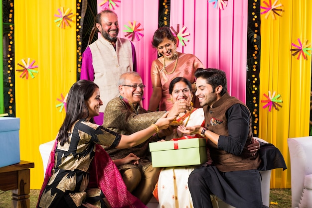 Famille Indienne Multigénérationnelle Mangeant Des Bonbons Tout En Célébrant Un Festival Ou Une Occasion Vêtue De Vêtements Traditionnels, Assise Sur Un Canapé Ou Un Canapé