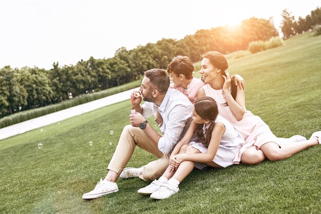 Famille de liaison de quatre personnes assises sur un champ herbeux soufflant des bulles