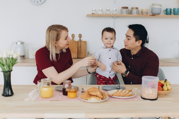 Famille multiculturelle heureuse. Papa asiatique et sa femme blonde caucasienne prennent le petit déjeuner avec leur beau fils dans la cuisine.