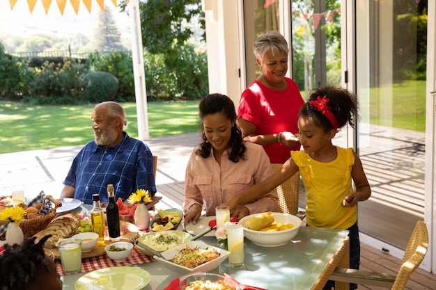 Une famille multiethnique et multigénérationnelle servant de la nourriture et s'asseyant à une table pour un repas dehors sur une terrasse au soleil, la petite-fille aidant sa grand-mère et plaçant des couverts sur la table