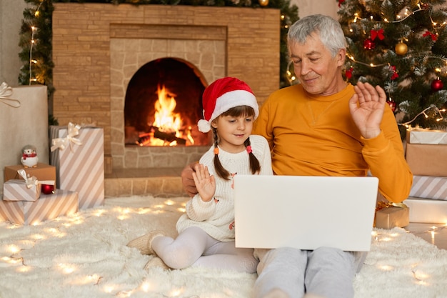 Famille de Noël, grand-père et petite-fille discutant sur internet avec un ordinateur portable assis sur le sol