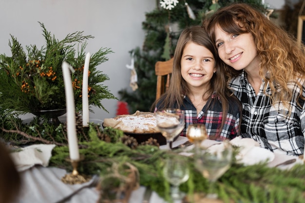 Famille à Noël à la table de fête Portrait d'une mère et d'une fille heureuses à une table décorée dans le salon Brindilles de bougies et un gros muffin de Noël pour le dîner Concept de vacances en famille