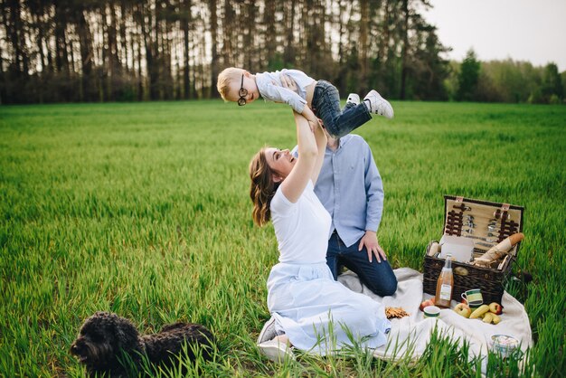 Famille en pique-nique dans la forêt. Dans le pré. Panier pique-nique avec de la nourriture. Le chien court et veut jouer. Un enfant avec des lunettes. Couleur bleue dans les vêtements. Pour manger dehors. Temps en famille.