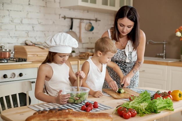 La famille prépare le déjeuner dans la cuisine. Maman apprend à sa fille et à son fils à préparer une salade de légumes frais. Alimentation naturelle saine, vitamines pour les enfants